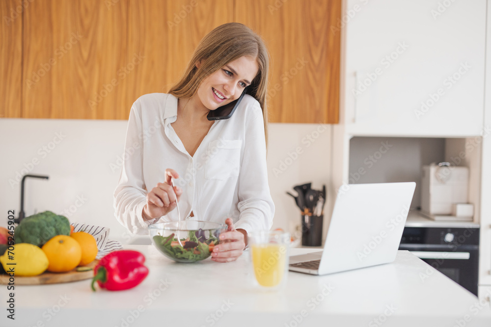 Happy and smiling blonde woman talking on the phone and looking for a recipe on the internet on a laptop in the kitchen Happy and smiling blonde woman talking on the phone and looking for a recipe on the internet on a laptop in the kitchen