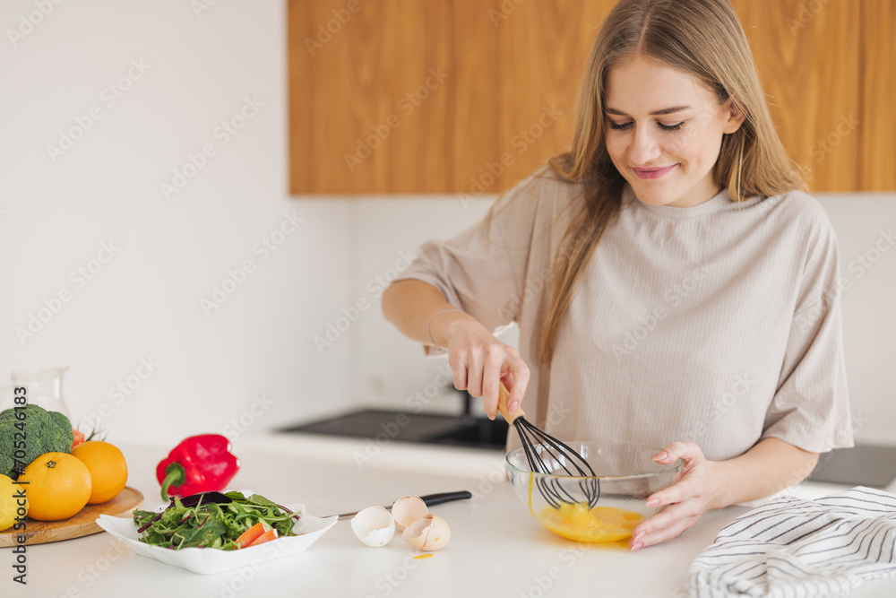 Happy pretty blonde woman in pajamas is beating eggs for breakfast in the kitchen at home. Happy pretty blonde woman in pajamas is beating eggs for breakfast in the kitchen at home.