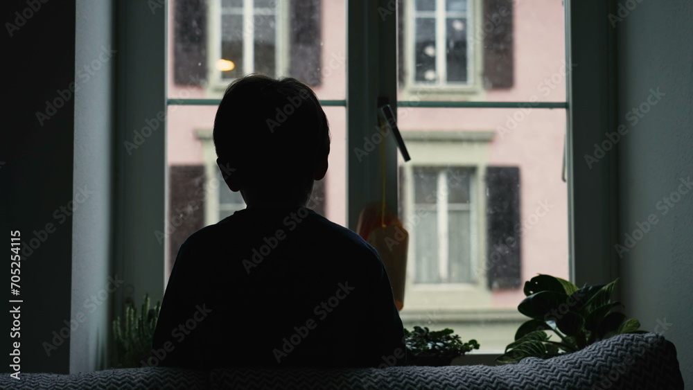 Silhouette of child staring at snow fall from home window during winter season. Back of young boy hypnotized by nature during moody day, December season Silhouette of child staring at snow fall from home window during winter season. Back of young boy hypnotized by nature during moody day, December season