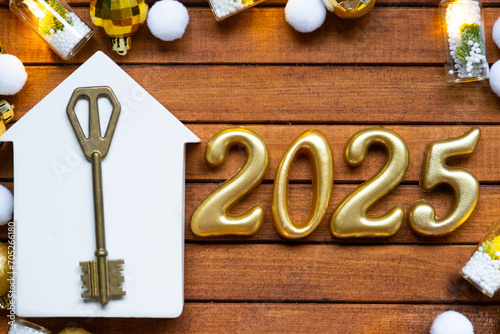 House key with tiny figure of home mock up on festive brown wooden background, lights of garlands. New Year 2025 wooden letters, greeting card. Purchase, construction, relocation, mortgage, insurance