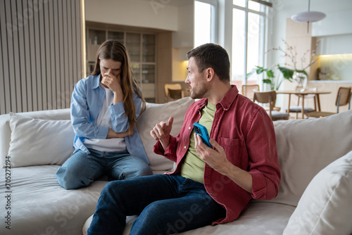 Angry irritated woman looking at man hiding smartphone display. Nervous wife listening explanations of sly justifying unfaithful husband who doesnt want to show mobile phone. Mistrust in family life photo