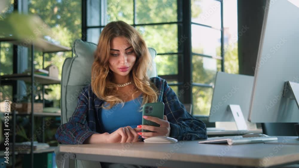 A smiling woman engages with smartphone in an office, green foliage from outside peeking through the large windows, creating a blend of work and nature