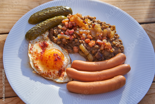 Close up picture on the white plate with traditional recipe from cuisine of Czech Republic. Lentils mash with roasted onion and pork sausages with sunny side up egg and sweet and sour pickled cucumber