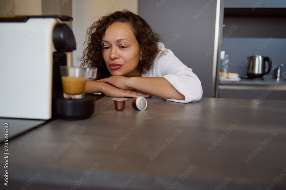 Sleepy young adult woman in white robe, admiring the capsule coffee machine making coffee for breakfast in the kitchen Sleepy young adult woman in white robe, admiring the capsule coffee machine making coffee for breakfast in the kitchen