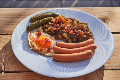 Simple but tasty dish from cuisine of Czech Republic. Small pork sausages served with lentil mash, sunny side up egg and pickled cucumbers on the white porcelain plate outside in the garden restaurant