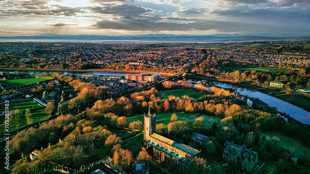 Aerial view of a cityscape at sunset with a prominent cathedral, lush green parks, and a river reflecting the warm sky in Lancaster.