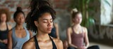 Young women in a diverse group practice meditation, sitting with closed eyes in the lotus position. They visualize together, cultivating self-awareness, serenity, and calmness in a yoga studio class.