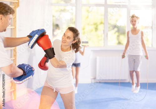 Positive woman wearing boxing gloves working out with husband during self-defense course, practicing punches on mitts while active tween daughter and son skipping rope. Sports family concept
