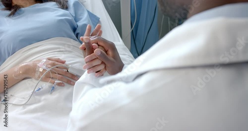 Diverse male doctor holding hand of female patient with oximeter in hospital bed, slow motion photo