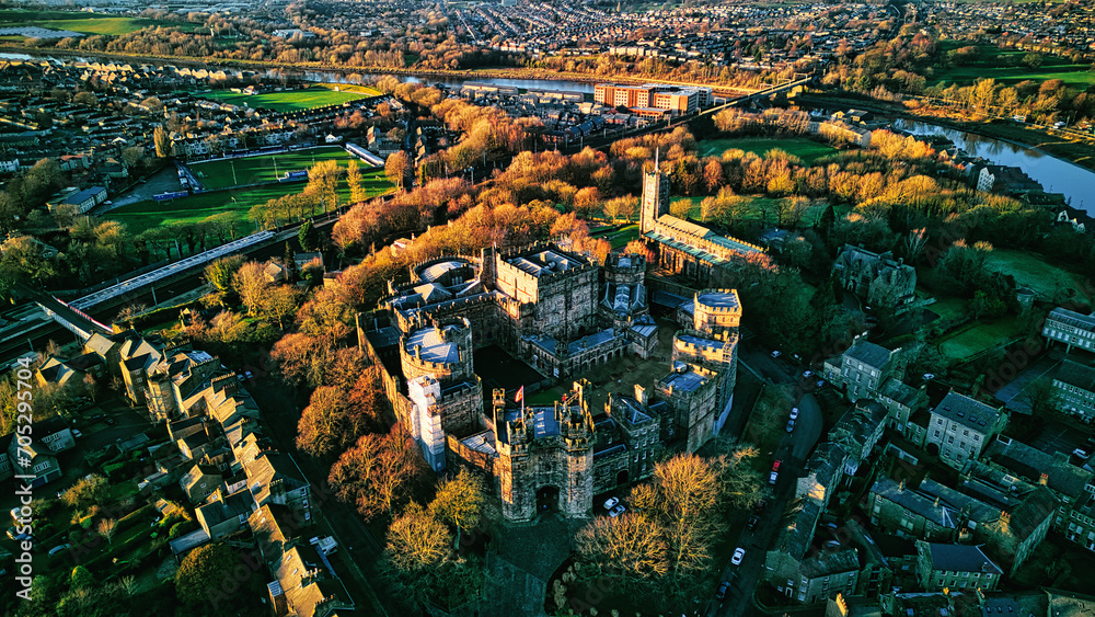 Aerial view of a historic Lancaster castle amidst a lush green landscape with surrounding urban area during golden hour.