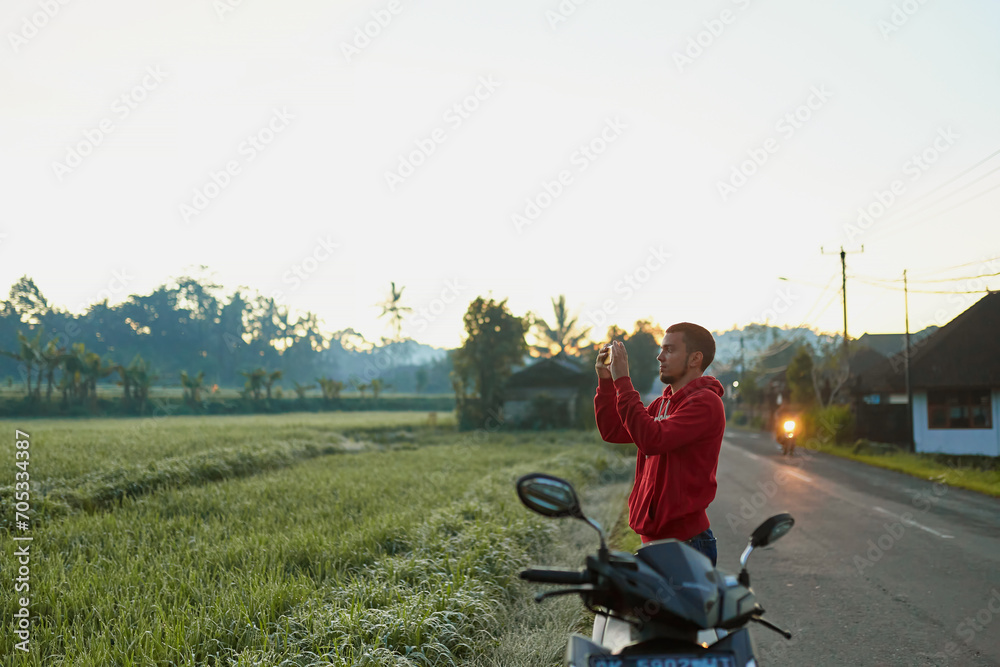 A young man stands on the side of the road near a rice paddy and takes a picture with his phone of Mount Agung on the popular tourist island of Bali. A young man stands on the side of the road near a rice paddy and takes a picture with his phone of Mount Agung on the popular tourist island of Bali.