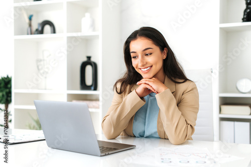 Amazing positive successful elegant brunette indian or arabian business woman, financial ceo, company employee, sitting at a work desk in the modern office, looking at laptop screen, smiling