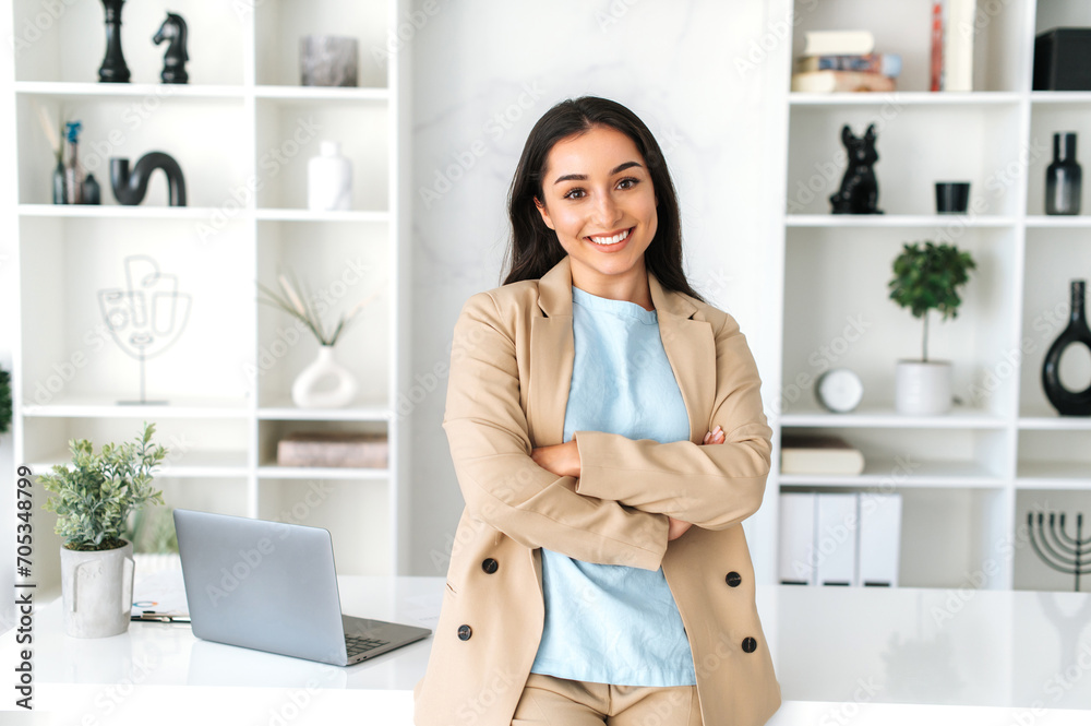 © Kateryna - Beautiful successful young adult indian or arabian business woman, ceo manager dressed in elegant suit standing in a modern office with arms crossed, friendly smiling at camera