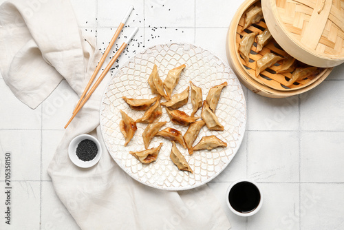 Plate with tasty Chinese jiaozi, sesame and bamboo steamer on white tile background