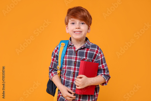 Happy schoolboy with backpack and book on orange background