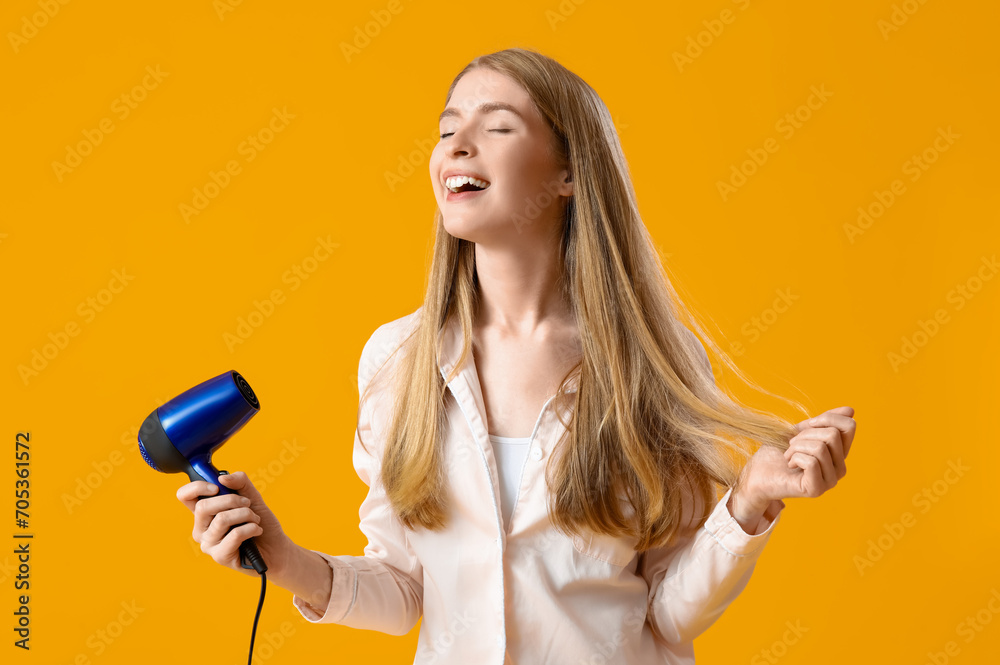 Young blonde woman in pajamas with hair dryer on yellow background Young blonde woman in pajamas with hair dryer on yellow background