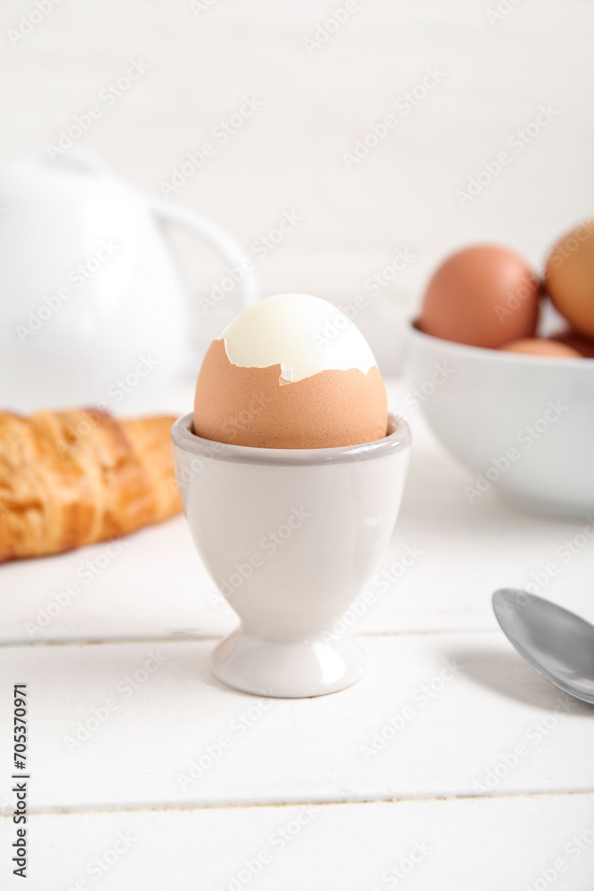 Holder with boiled chicken egg, teapot and croissant on white wooden background Holder with boiled chicken egg, teapot and croissant on white wooden background