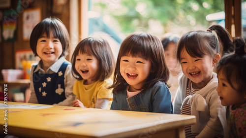 日本の幼稚園児5人が私服で机の周りに座って笑っている写真、背景の大きな窓から緑が見える木でできた壁の室内