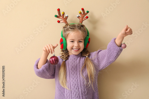 Happy little girl with Christmas baubles  reindeer antlers and earmuffs on beige background