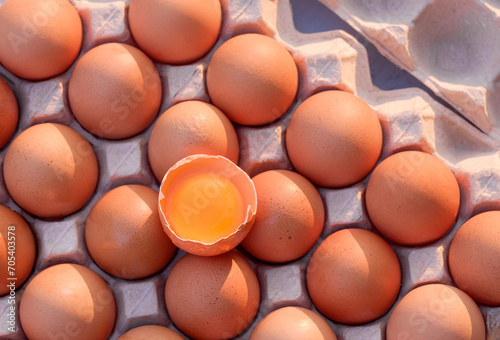 Flat lay of one cracked egg with yolk on top of fresh brown chicken eggs in carton tray, top view raw organic food background with copy space 