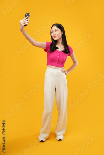 A photo of a young Asian woman wearing a casual pink t-shirt taking a selfie with a smartphone cellphone in her hands. Isolated on a yellow background. 