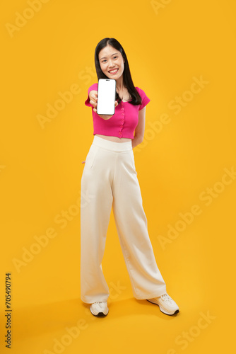 Portrait of a happy young Asian lady showing a smartphone with a blank screen, smiling and isolated on a yellow background. Advertise concept. 