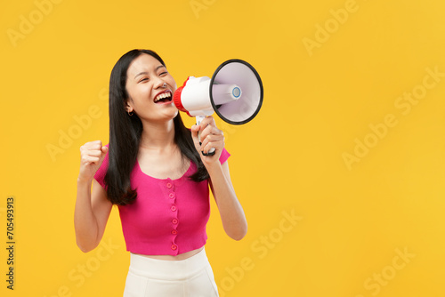 Photo of a young Asian woman holding a megaphone surprised with a happy face, wearing a pink t-shirt, and isolated on a yellow background.