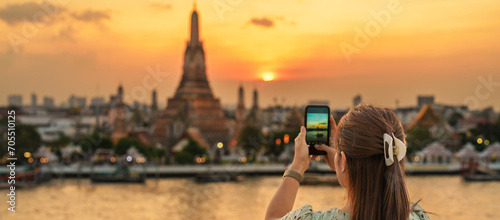 tourist woman enjoys view to Wat Arun Temple in sunset, Traveler take photo to Temple of Dawn by smartphone from rooftop bar. Landmark and Travel destination in Bangkok, Thailand and Southeast Asia photo