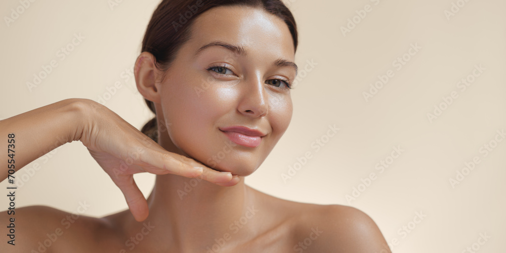 Happy beautiful young adult woman touching her perfect face with healthy shiny skin, hand on chin. Beauty photo of gorgeous girl after morning beauty cosmetic routine finishes daily makeup on beige - obrazy, fototapety, plakaty 