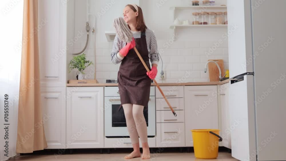 Smiling joyful woman in apron making domestic work washing floor in modern interior kitchen being in good mood holding mop as microphone singing and having fun while cleaning house Smiling joyful woman in apron making domestic work washing floor in modern interior kitchen being in good mood holding mop as microphone singing and having fun while cleaning house