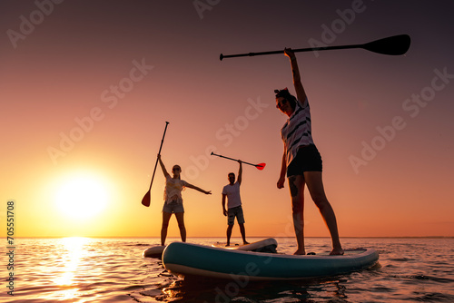 Three happy frieSilhouettes of three friends are standing in calm lake with sup boards in handsnds are paddleboarding on sups at sunset