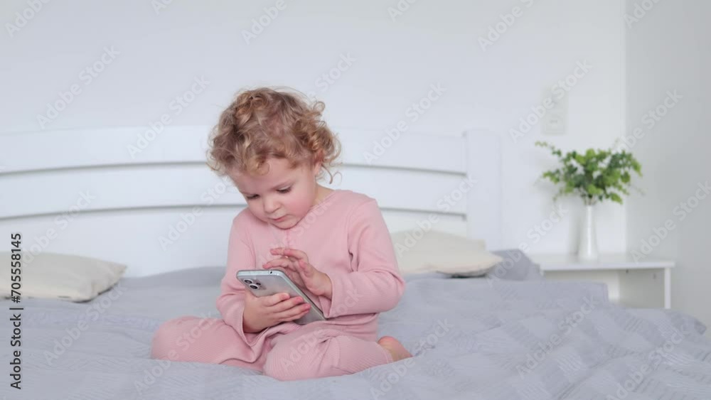 Little toddler wavy haired blond girl sitting on bed and using smartphone choosing cartoons for watching or kids game for playing while having gadget time Little toddler wavy haired blond girl sitting on bed and using smartphone choosing cartoons for watching or kids game for playing while having gadget time