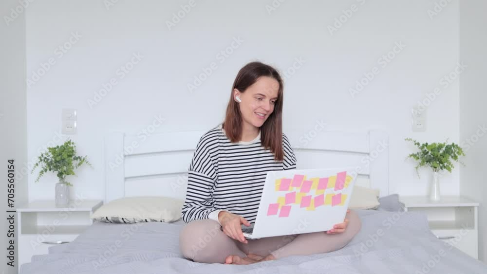 Woman relaxing on bed using laptop computer for video call sitting in her cozy bedroom in earphones waving hand hello gestured greeting her friend enjoying online conversation Woman relaxing on bed using laptop computer for video call sitting in her cozy bedroom in earphones waving hand hello gestured greeting her friend enjoying online conversation