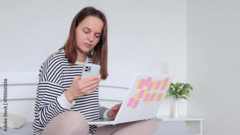 Concentrated focused woman sitting on bed in cozy bedroom working on phone and laptop at home wearing casual striped shirt doing her remote job from home office Concentrated focused woman sitting on bed in cozy bedroom working on phone and laptop at home wearing casual striped shirt doing her remote job from home office