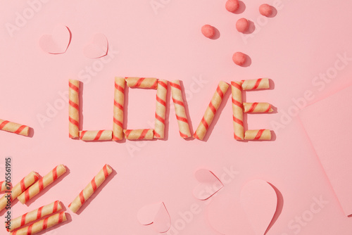 The word LOVE is arranged from wafer roll on a pink background with paper hearts and strawberry candy balls. Ideal space for Valentine's Day. Flat lay.