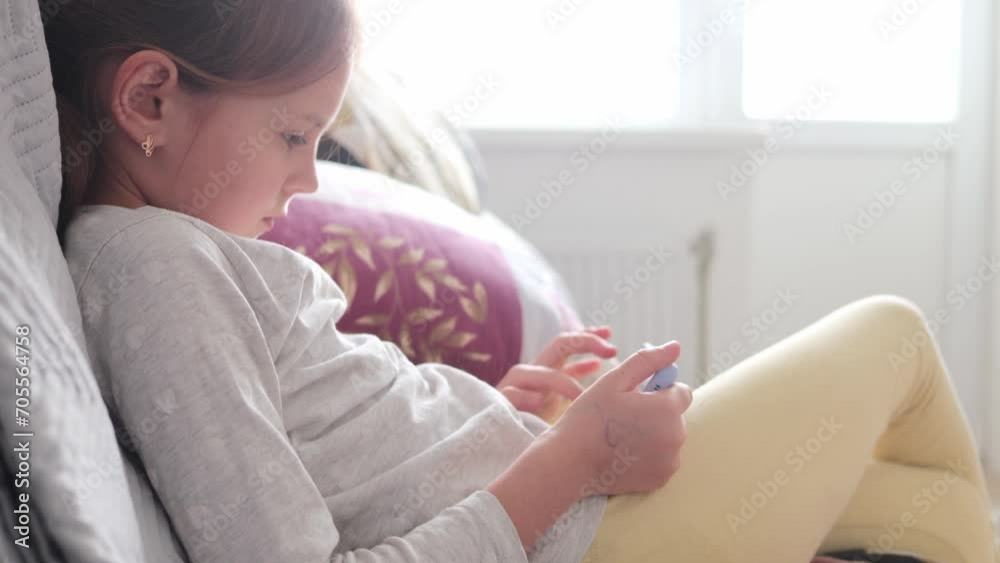 Cute calm concentrated little girl using technology device sitting on sofa playing on phone watching video browsing internet web pages scrolling online