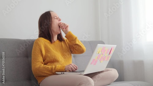 Exhausted tired sleepy woman freelancer working online on her laptop typing on keyboard being sleepless yawning while doing her remote job sitting on sofa in living room photo