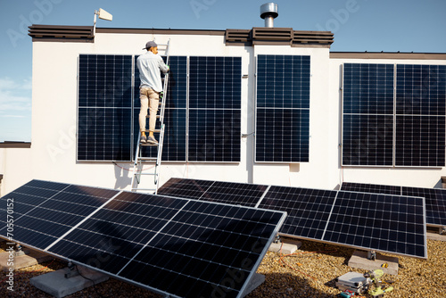 Man installing solar panels, climbing on a ladder on the roof of his house. Wide angle view on a roof with solar power station. Renewable energy and sustainability concept