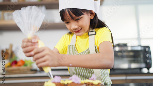 Little toddler asian girl child in apron and chef hat whipped cream decorating preparing homemade cupcakes in home kitchen. A Little girl preparing and decorating homemade cake. Children cooking