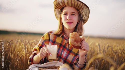 Young girl farmer in sun hat eats loaf of bread biting it drinking milk sits on wheat field with grain bag on knees. Harvesting, summer on farm, food business, agribusiness, woman worker has break. photo