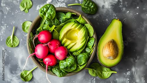 A bowl of fresh vegetables and greens on a black textured surface. Inside the bowl are bright red radishes with green stems attached, vibrant green spinach leaves, and slices of ripe avocado.