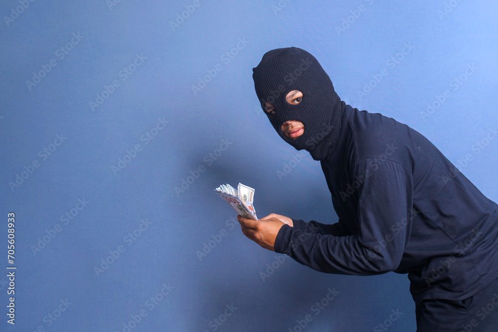 Bank robber wearing balaclava holding bunch of stollen dollars, isolated on blue studio background - obrazy, fototapety, plakaty 
