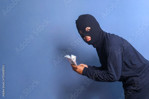 Bank robber wearing balaclava holding bunch of stollen dollars, isolated on blue studio background photo