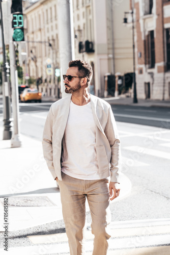 Handsome confident stylish hipster lambersexual model. Sexy modern man dressed in elegant white suit jacket. Fashion male posing in the street background in Europe city at sunset. In sunglasses