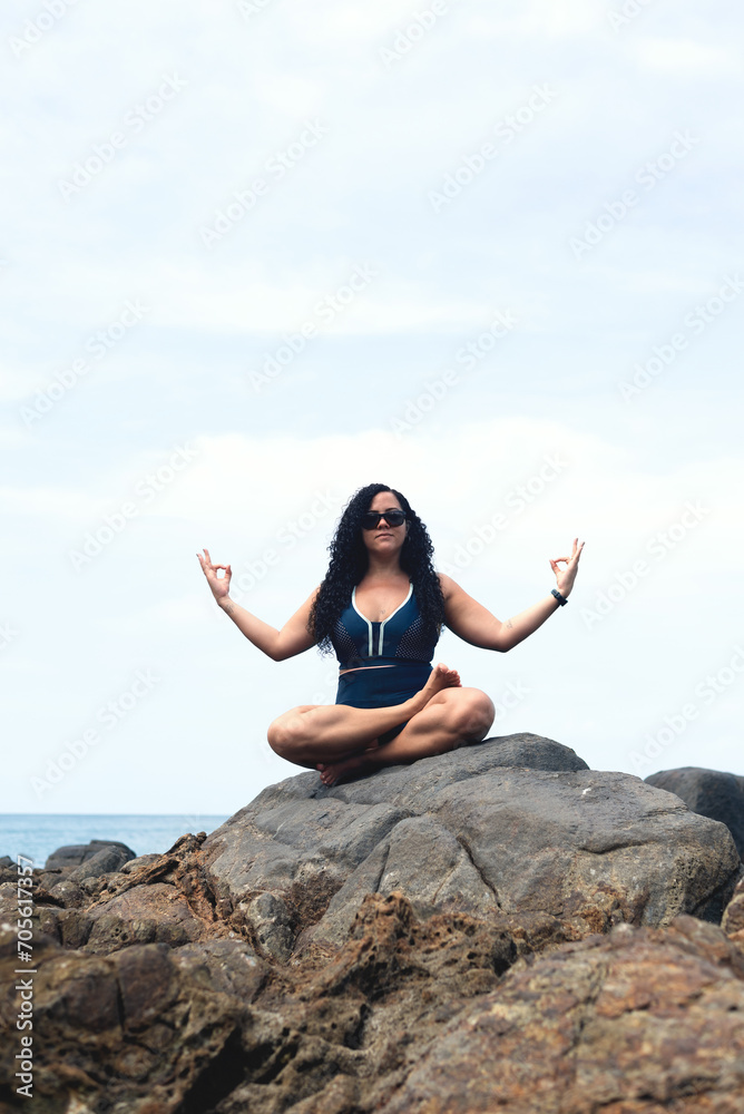 Beautiful, curly-haired woman sitting on the rock of a beach doing yoga exercise. Beautiful, curly-haired woman sitting on the rock of a beach doing yoga exercise.