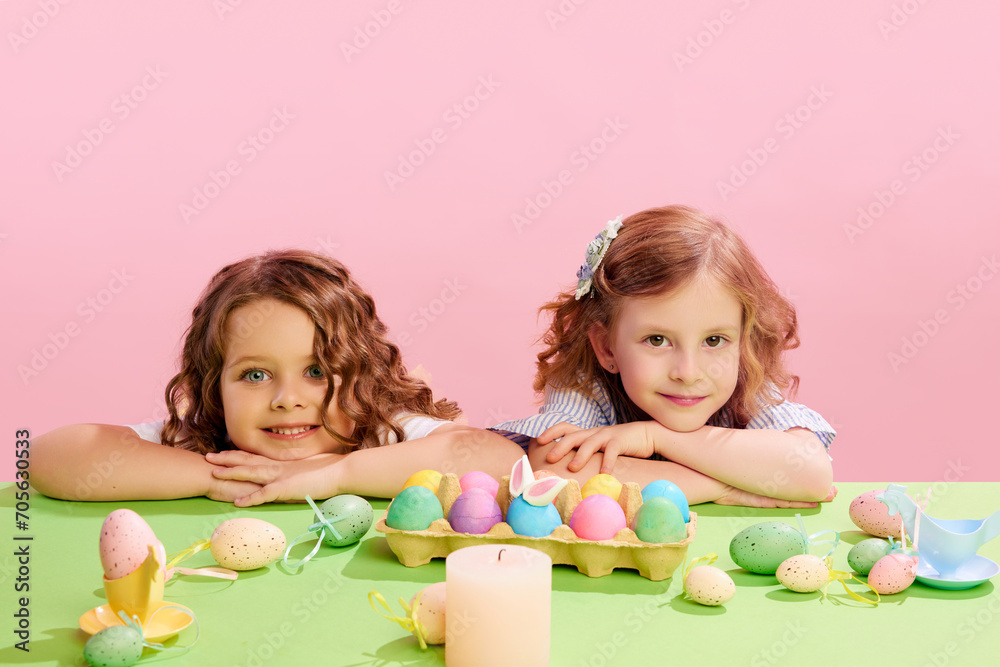 Portrait of beautiful little girls lying on table with decorated, painted eggs against pink background. Concept of Easter holiday, celebration, traditions, childhood, happiness Portrait of beautiful little girls lying on table with decorated, painted eggs against pink background. Concept of Easter holiday, celebration, traditions, childhood, happiness