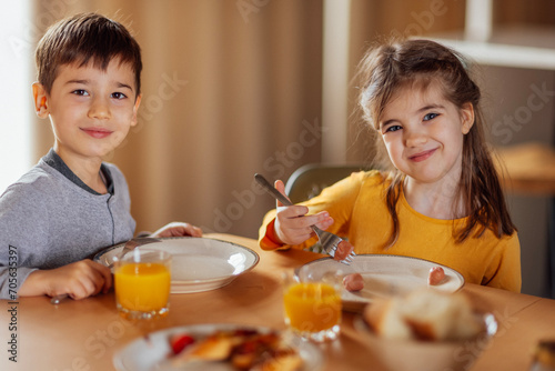 Two small children are sitting at dining table and having breakfast. Cute kids smile and look at camera.