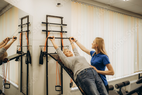 Positive senior woman doing stretching exercises on Pilates reformer as part of injury rehabilitation course. Therapeutic physical training concept photo