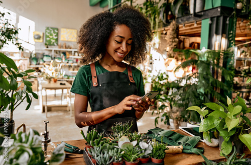 Beautiful african american woman florist in a green t-shirt and apron stands in the shop with plants on the table and scrolls the phone while working. Busy business lady taking orders online. photo