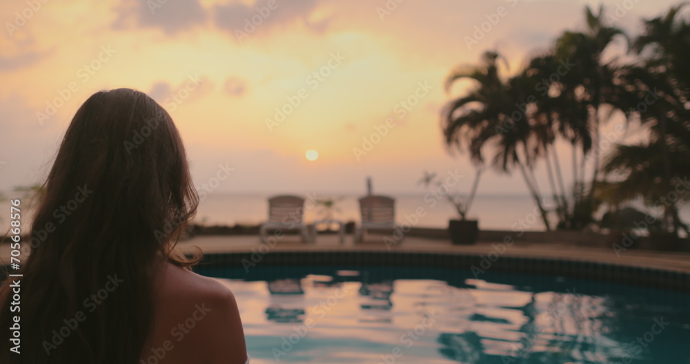 Long hair brunette enjoy bright colorful sunset on tropical island. Orange sky reflected in swimming pool, palm trees and deck chairs. Lifestyle travel on summer holiday vacation. Back view